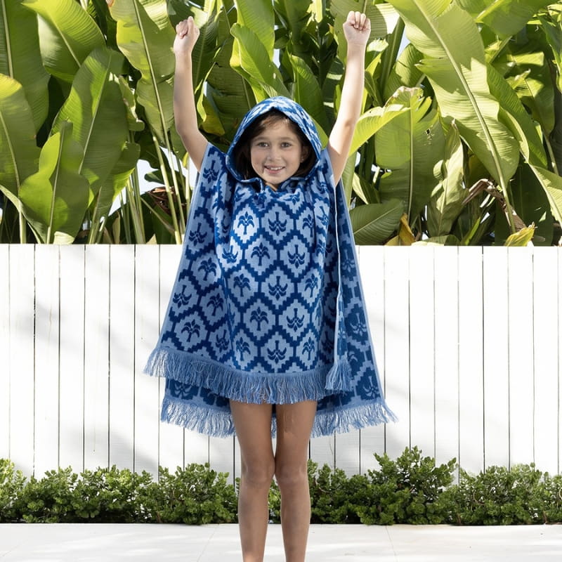 alt="A kid wearing a blue poncho featuring a stylish palm tree pattern with knotted tassels along the ends"