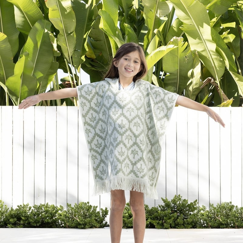 alt="A kid wearing a sage green poncho featuring a stylish palm tree pattern with knotted tassels along the ends"