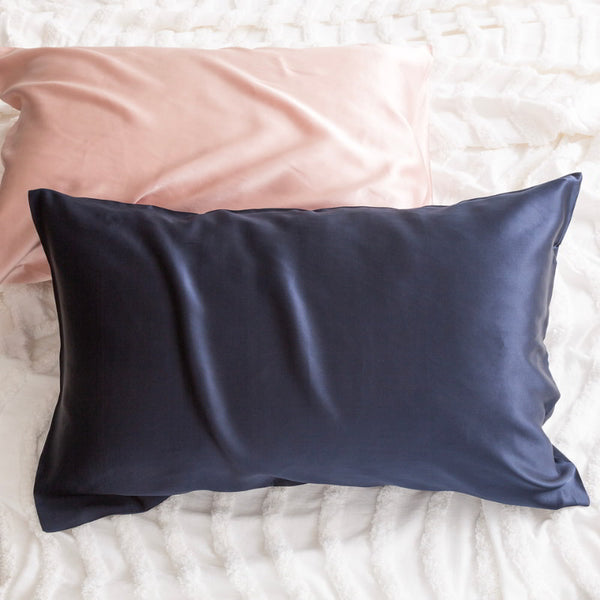 alt="Pink and navy pillowcase on a white bed. Crafted from 100% pure mulberry silk, these luxurious pillows offer a refreshing experience for your skin. "