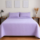 A neatly made bed features the Morgan and Reid Mosaic Lilac Coverlet Set with matching pillows, wooden nightstands, minimalist art above, and a vase of dried grass. The lightweight coverlet from Morgan and Reid is machine washable for easy care.