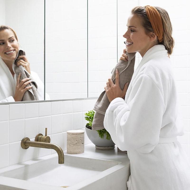 alt="A woman wearing a white microplush robe, looking at herself in the mirror"