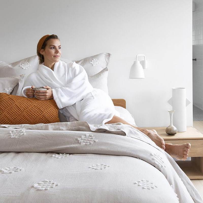 alt="A woman wearing a microplush white robe drinking coffee while sitting on her bed"