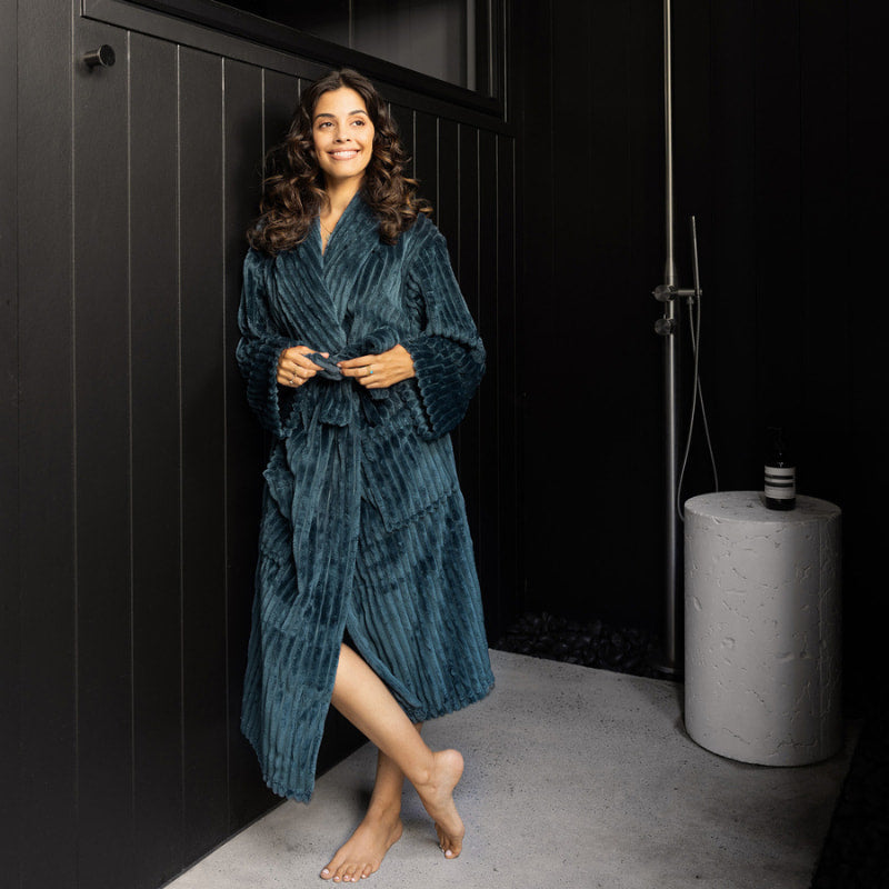 A woman draped in a sumptuous blue velvet robe stands in front of a bold black wall, radiating elegance and refinement.