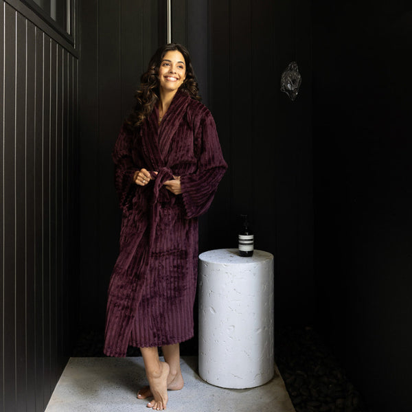 A woman draped in a sumptuous plum velvet robe stands in front of a bold black wall, radiating elegance and refinement.