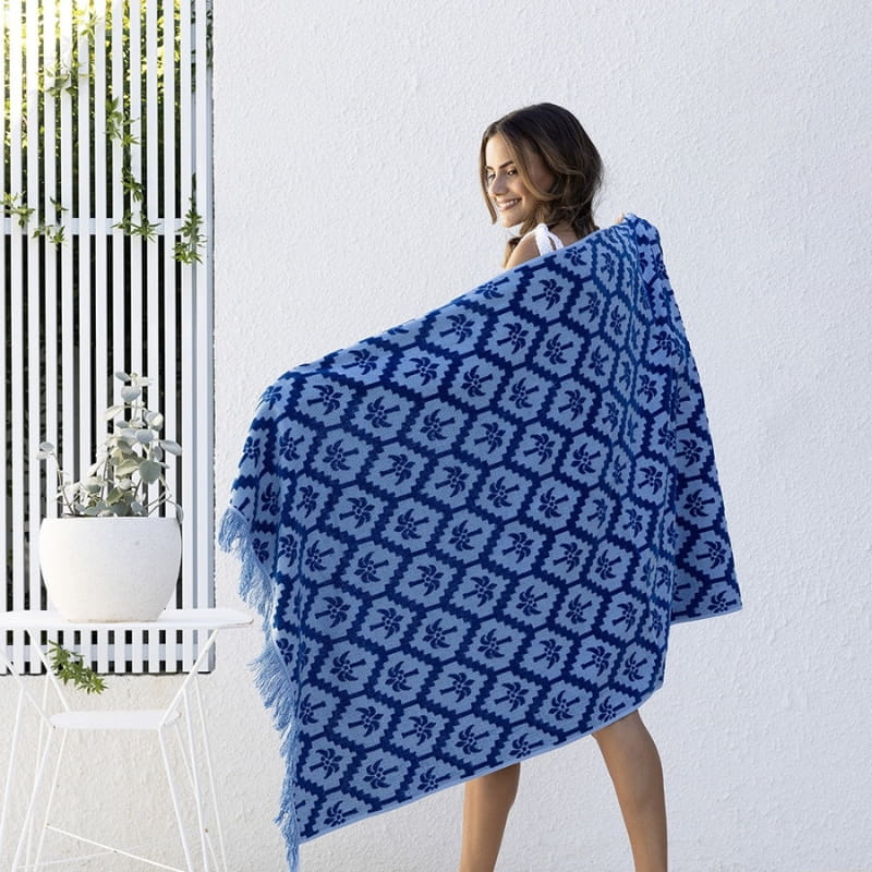 alt="A woman holding a cobalt blue beach towel designed with palm tree pattern and tassels on both ends"