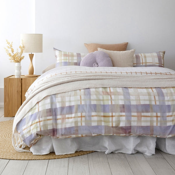 alt="A digitally printed hand-painted watercolour check pattern in sage green, lilac ash, and tan on a crisp white base quilt cover set in a cosy bedroom"