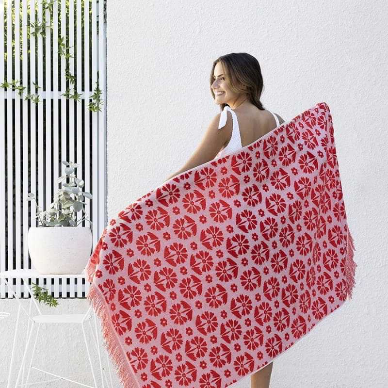 alt="A woman holding a red beach towel designed with floral pattern and tassels on both ends"