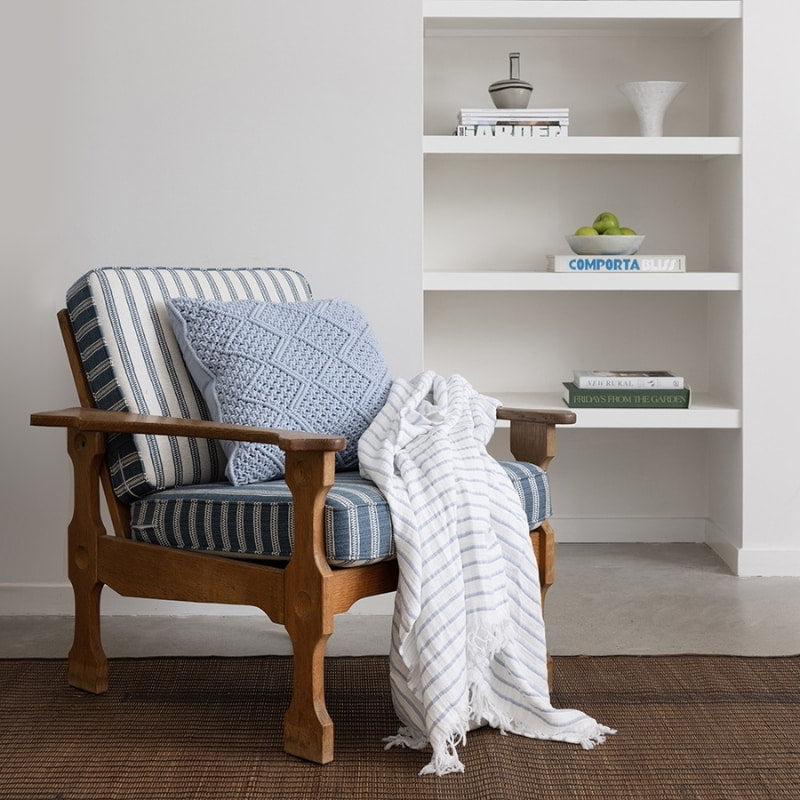 alt="Striped white and blue throw with fringe at both ends placed on a wooden chair in a coastal-theme living space"