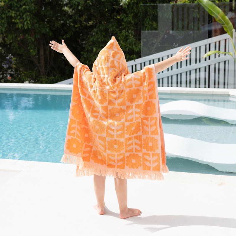 A kid wrapped in an HiLo Bloom orange poncho by the pool, ready for post-swim relaxation and sun protection.