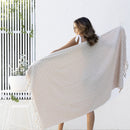 alt="A woman holding a stylish natural beach towel with a small check pattern achieved through a series of high and low sections of towelling finished with a beautiful fringe along the ends"