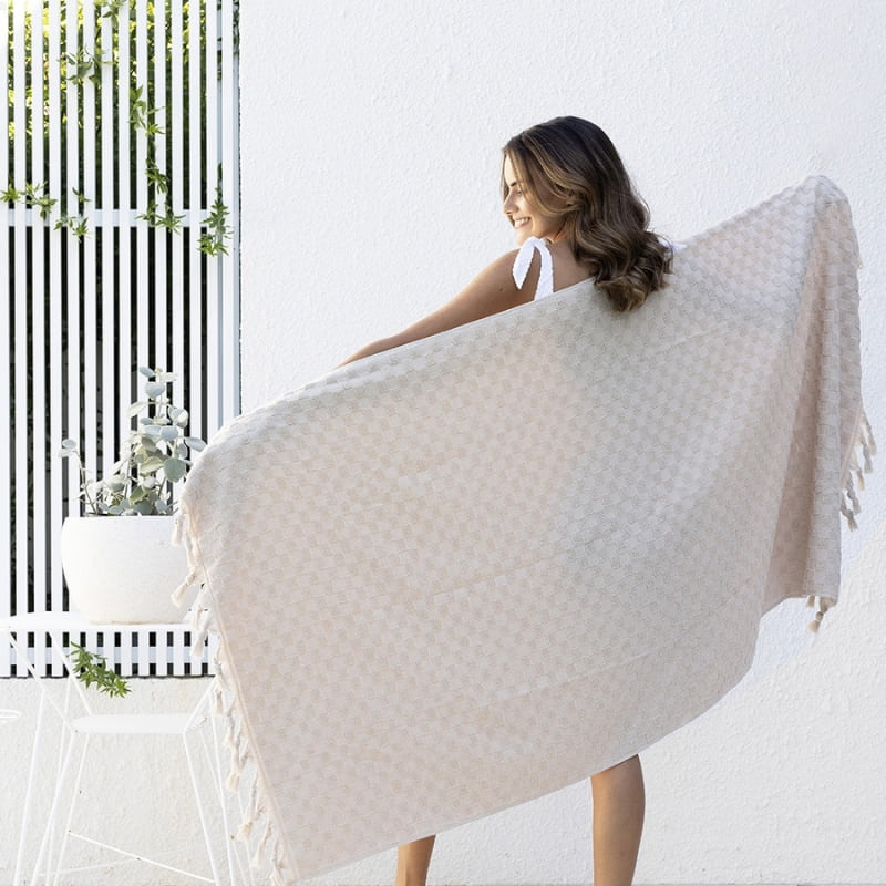 alt="A woman holding a stylish natural beach towel with a small check pattern achieved through a series of high and low sections of towelling finished with a beautiful fringe along the ends"