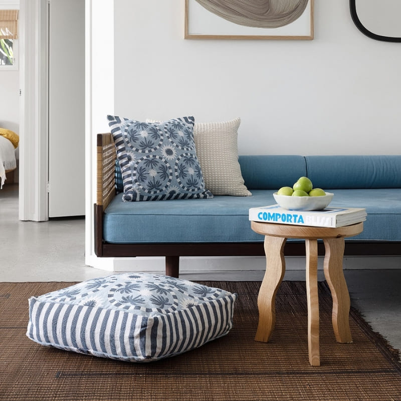 alt="A light blue and dark blue designed with palm tree pattern square cushion placed on a sofa in a cosy room"