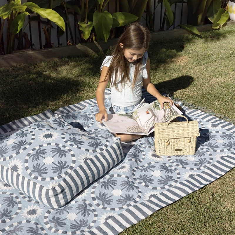 alt="A girl sitting on a light blue and dark blue designed with palm tree pattern floor cushion in an outdoor setting"
