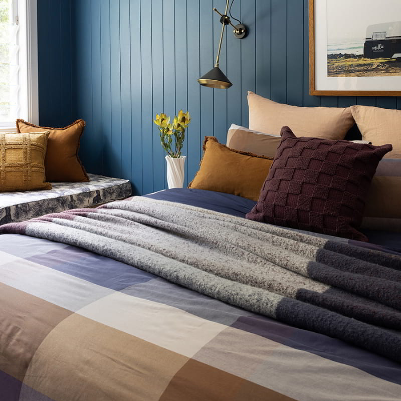 A cosy bedroom with blue walls and a bed featuring a plaid set in purple, navy, and beige tones.