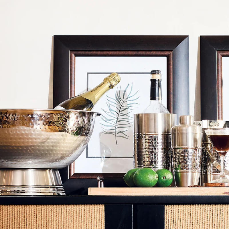 alt="An elegant hammered chrome champagne bowl with a chilled bottle of champagne inside, surrounded by a curated collection of barware and glassware."