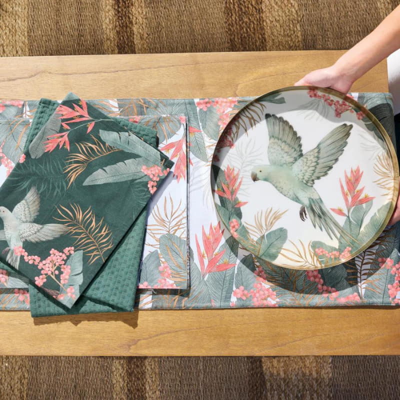 alt="A round serving tray and placemats with a tropical design featuring a flying bird amidst lush foliage"