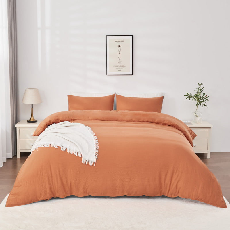 alt="A rust-coloured quilt cover set on a bed with white fringed throw in a cosy bedroom"