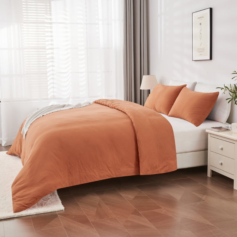 alt="Side view of a rust-coloured quilt cover set on a bed with white fringed throw in a cosy bedroom"