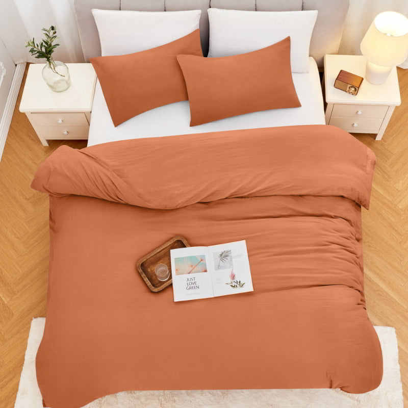 alt="Top view of a rust-coloured quilt cover set on a bed with white fringed throw in a cosy bedroom"