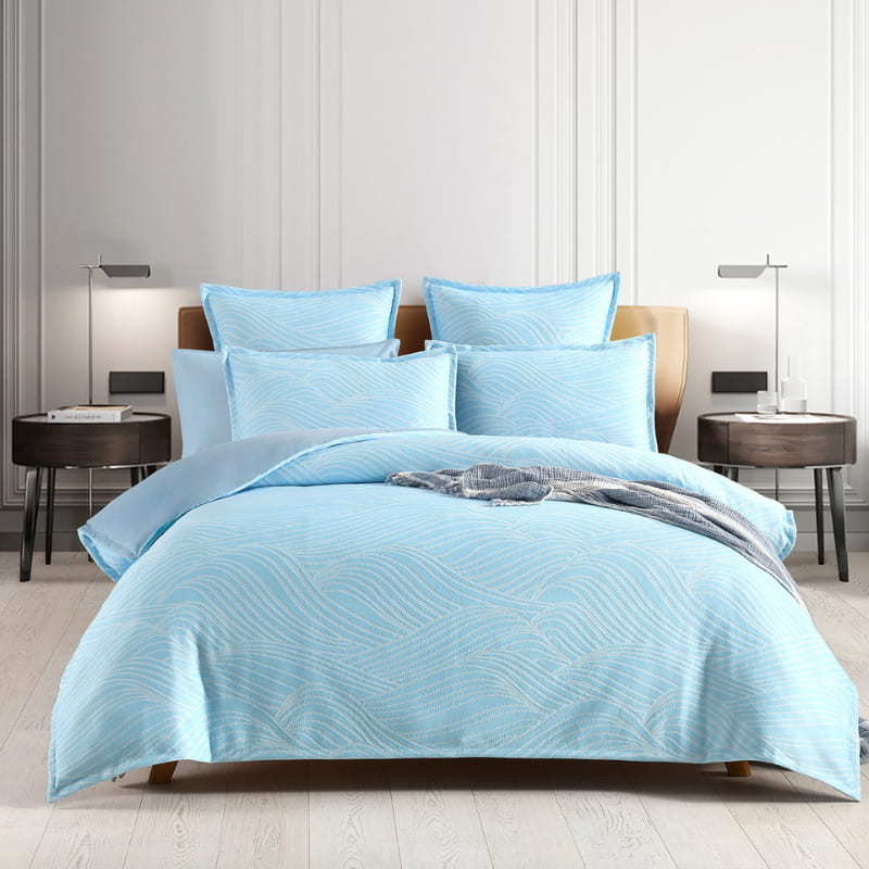 alt="Showcasing a shades of blue bedding set with a beach wave design in a cosy bedroom."