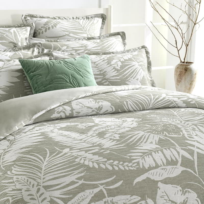 alt="Showcasing a shade of sage green quilt cover featuring intricate island themes with swaying palm trees in a tropical rainforest in a luxurious bedroom."