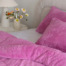 Close-up of pink textured pillows on a bed with a lamp and flowers in the background.