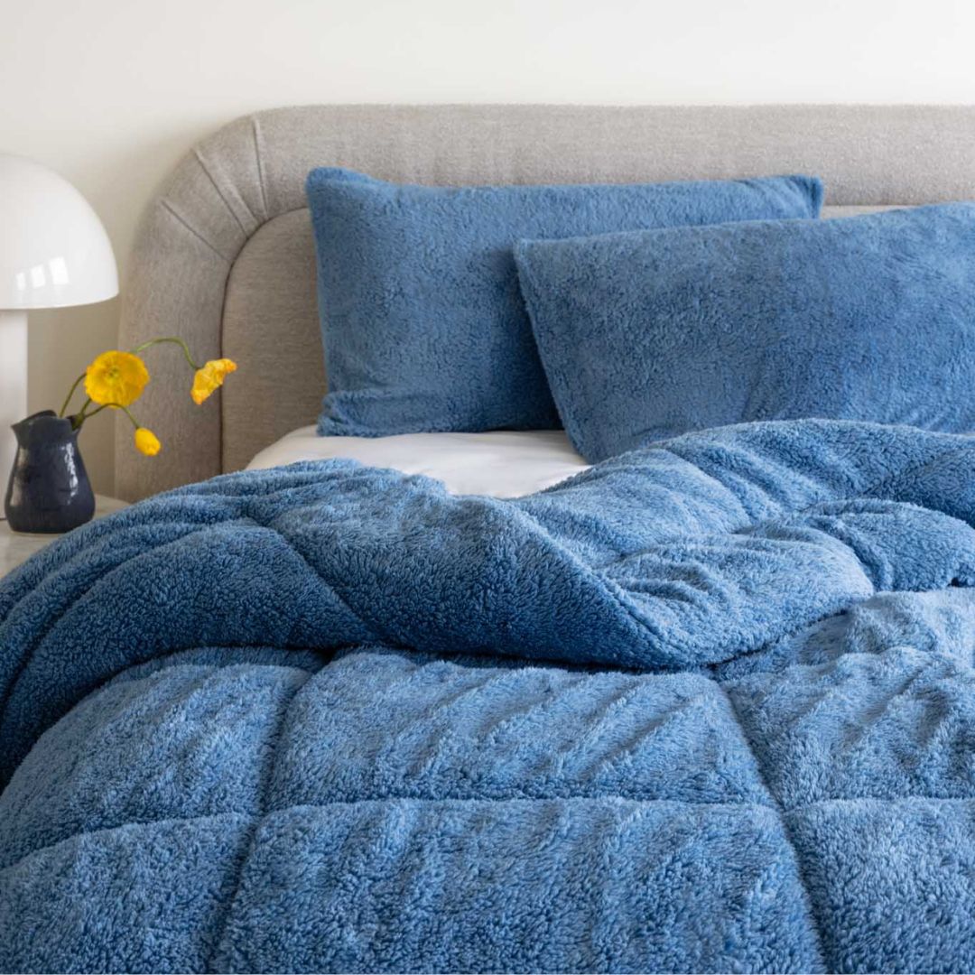 Blue textured comforter on a bed with pillows and a vase of yellow flowers.
