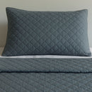 A neatly made bed features the Morgan and Reid Argyle Shade Coverlet Set in dark gray with matching pillows, creating a contemporary vibe. Above, three abstract art pieces hang, while a wooden table with red flowers sits to the right.