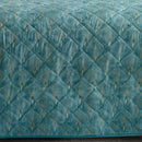 A bright bedroom features the Morgan and Reid Art Deco Coverlet Set in teal blue, with a quilted microfibre bedspread and matching pillows by Morgan and Reid, styled with a wooden headboard, bedside tables, lamp, and a window reflecting autumn trees.