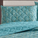 A bright bedroom features the Morgan and Reid Art Deco Coverlet Set in teal blue, with a quilted microfibre bedspread and matching pillows by Morgan and Reid, styled with a wooden headboard, bedside tables, lamp, and a window reflecting autumn trees.