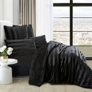 A modern bedroom features the Morgan and Reid Black Velvet Stripe Snuggle Fleece Comforter Set on a neatly made bed with striped pillows, neutral walls, abstract art, a beige chair, round black side table with white flowers, and a lamp.
