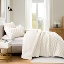 A neatly made bed showcases the Morgan and Reid Checkered Cream Snuggle Fleece Comforter Set with textured pillows, against a light wall, paired with a wooden headboard, bedside table, lamp, and a large leafy plant.