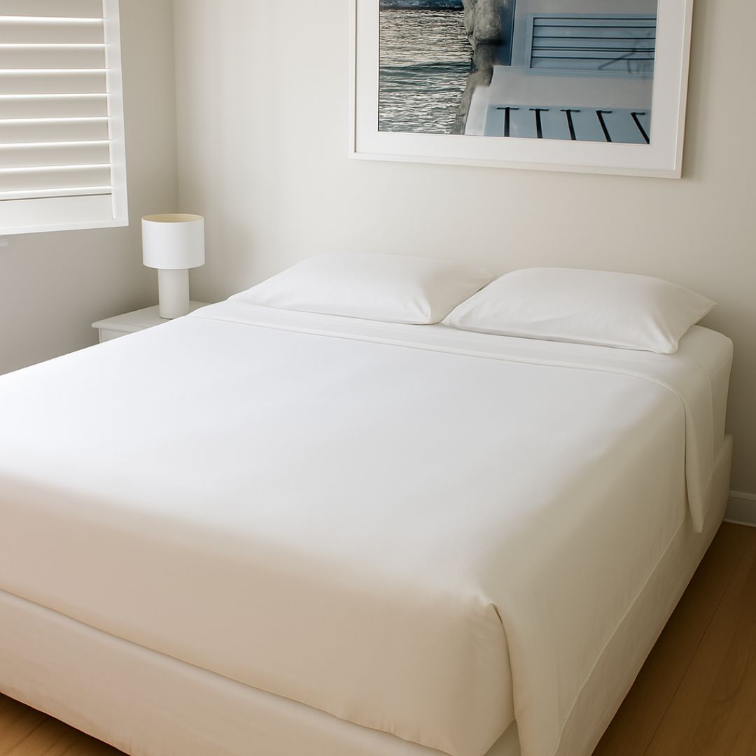 A bright, minimalist bedroom with a neatly made bed dressed in the Morgan and Reid Ultra Cooling Sheet Set. White sheets and pillows complement a bedside lamp and framed photo. A label reads “Made For Hot Sleepers” by Morgan and Reid.