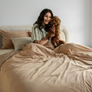 Woman sitting on a bed holding a dog, surrounded by beige bedding and pillows.