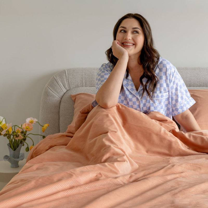 Woman in a checkered shirt sitting on a bed with peach-colored bedding, smiling.