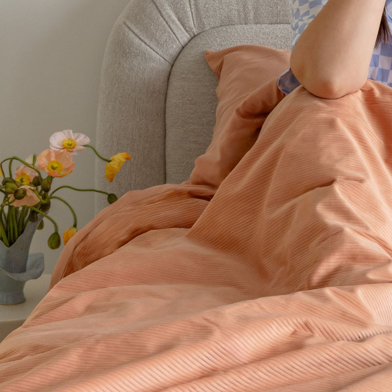 Person wrapped in a peach-colored blanket on a couch with a vase of flowers on a table.