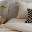 Close-up of a bed with beige pillows and a checkered pillow, wooden nightstand with plant, and gray upholstered headboard.