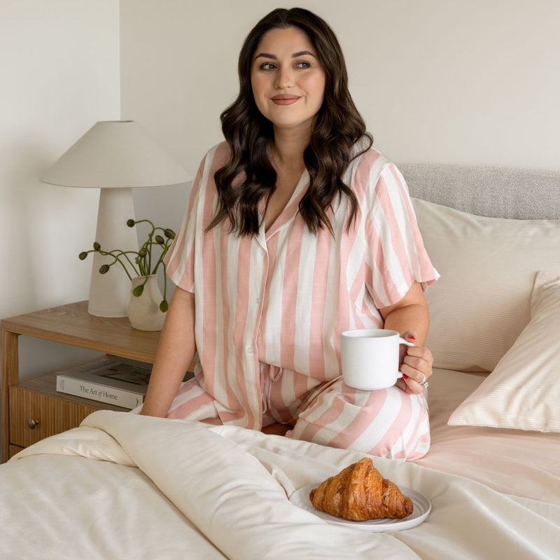 Woman in pink and white striped pajamas holding a mug on a bed with a plate of croissants.
