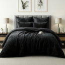 A neatly made bed features the Morgan and Reid Black Velvet Crushed Snuggle Fleece Comforter Set, paired with pillows and flanked by nightstands with lamps. Two botanical prints hang above the bed in a light-walled, carpeted room. A cup and saucer rest on the bed.