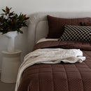 Bed with brown bedding and pillows, next to a plant on a side table.