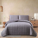 Gray quilted bedding set on a bed in a bedroom with a beige wall and wooden nightstand.