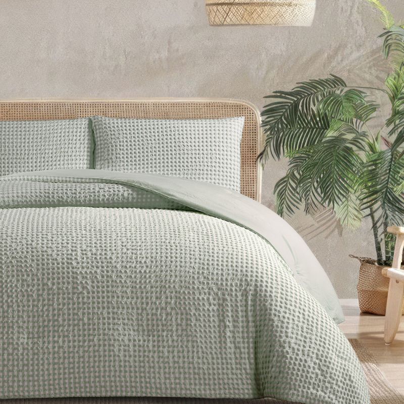 A calming bedroom displays the Morgan and Reid Eucalyptus Gingham Comforter Set on a neatly styled bed with matching pillowcases, rattan furniture, natural pendant lamp, indoor plant, and a woven rug.