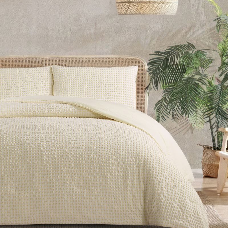 A modern bedroom features the Morgan and Reid Lemon Butter Gingham Comforter Set on a neatly made bed with matching pillowcases, rattan bedhead, neutral walls, woven pendant light, and a wooden side table with decorative accents.