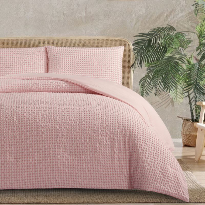 Pink textured bedspread on a bed with a wicker headboard, plants in the background.