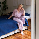 A neatly made bed features the Morgan and Reid Honeycomb Midnight Coverlet Set with matching pillows in a minimalist bedroom, complemented by a wooden nightstand, a white vase of red flowers, and three line art prints above the bed.