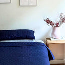 A neatly made bed features the Morgan and Reid Honeycomb Midnight Coverlet Set with matching pillows in a minimalist bedroom, complemented by a wooden nightstand, a white vase of red flowers, and three line art prints above the bed.