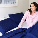 A neatly made bed features the Morgan and Reid Honeycomb Midnight Coverlet Set with matching pillows in a minimalist bedroom, complemented by a wooden nightstand, a white vase of red flowers, and three line art prints above the bed.