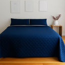 A neatly made bed features the Morgan and Reid Honeycomb Midnight Coverlet Set with matching pillows in a minimalist bedroom, complemented by a wooden nightstand, a white vase of red flowers, and three line art prints above the bed.