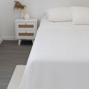 A neatly made bed is dressed in the Morgan and Reid Heavy-Weight Crisp Cotton Sheet Set, with pillows beside a white two-drawer nightstand holding vases with dried grasses. The gray wood floor shows part of a white rug.