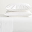 A neatly made bed is dressed in the Morgan and Reid Heavy-Weight Crisp Cotton Sheet Set, with pillows beside a white two-drawer nightstand holding vases with dried grasses. The gray wood floor shows part of a white rug.